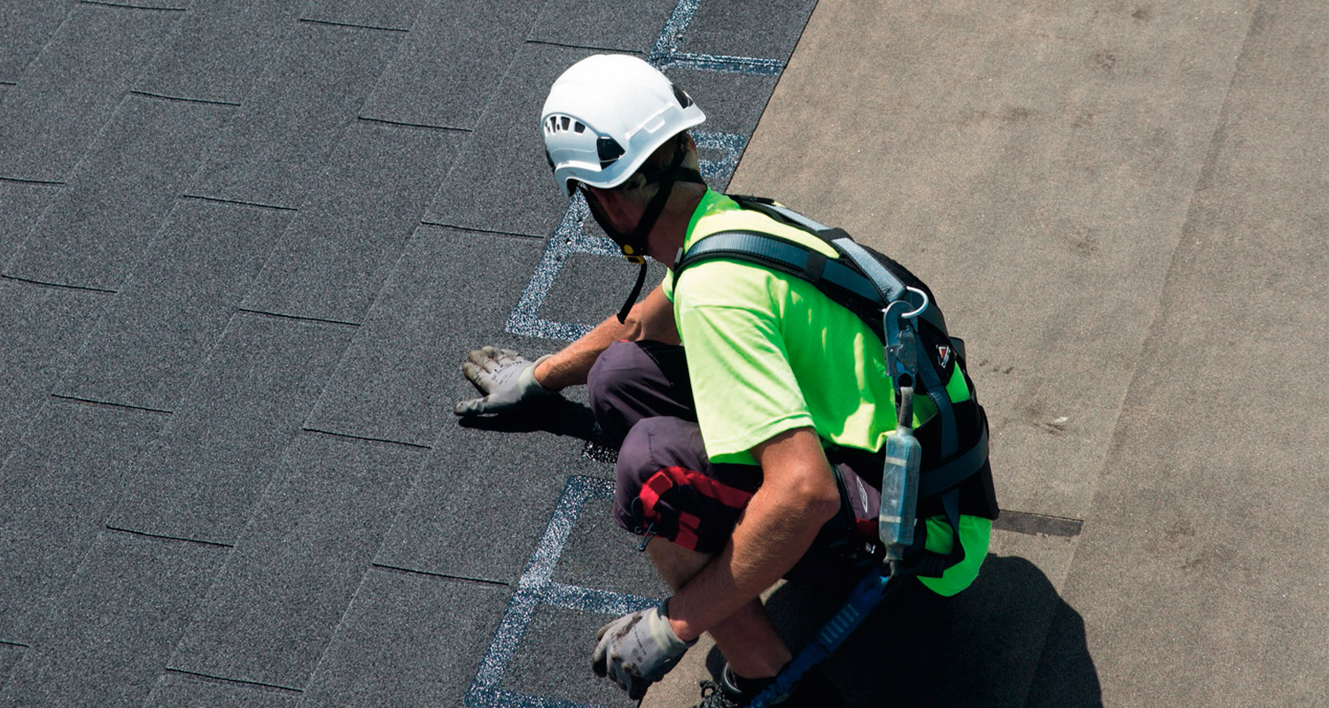 Kattoasentaja neonkeltaisessa paidassa, valkea kypärä ja turvavaljaat, asentaa harmaita huopia.