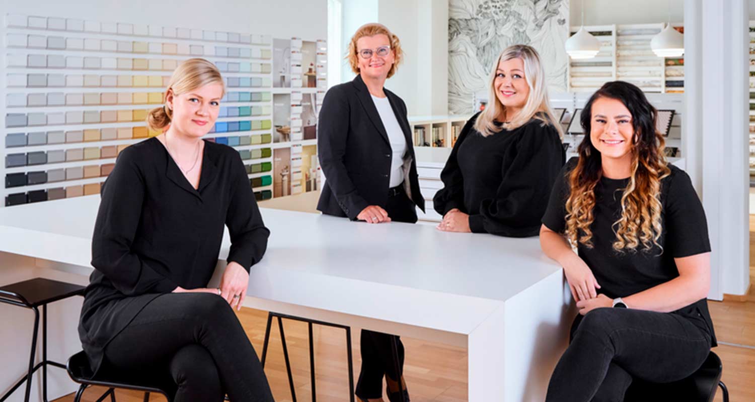 Neljä mustiin pukeutunutta naista valkoisen pöydän äärellä värikarttaseinän edessä.