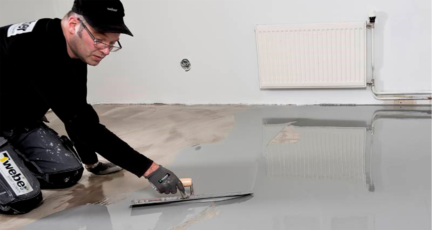 Mies levittää harmaata lattiatasoitetta viistetyökalulla valkoisen seinän vieressä.