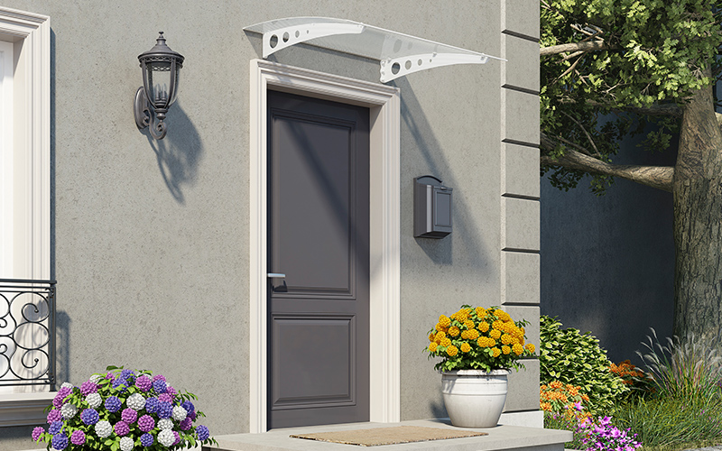 Harmaan talon sisäänkäynti: tumma ovi, lasikatto, postilaatikko ja lilaisia sekä keltaisia kukkia.