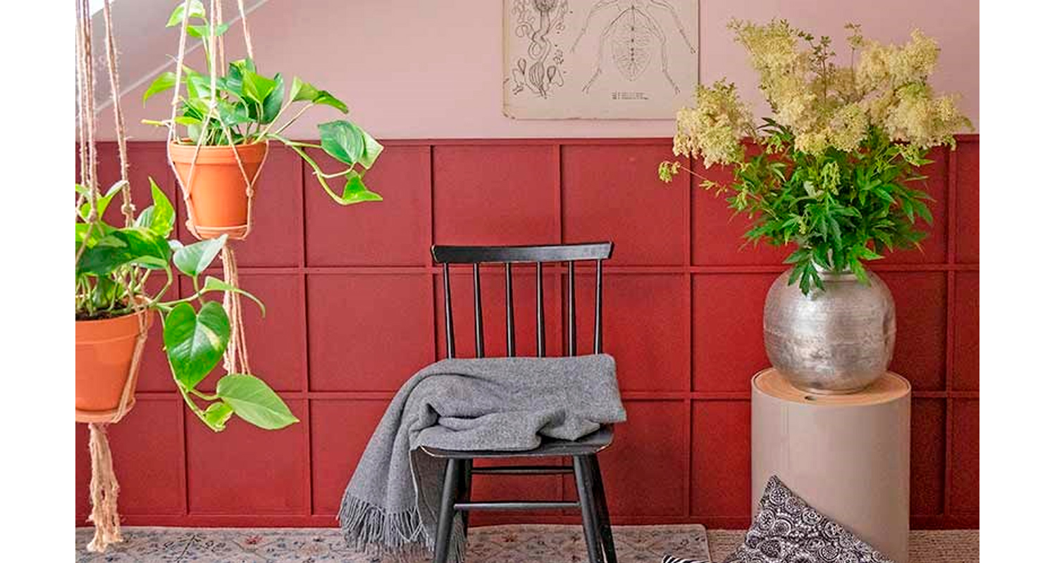 Tuoli harmaalla viltillä punaisella taustalla, hopeamaljakko kukkineen ja roikkuvia viherkasveja.