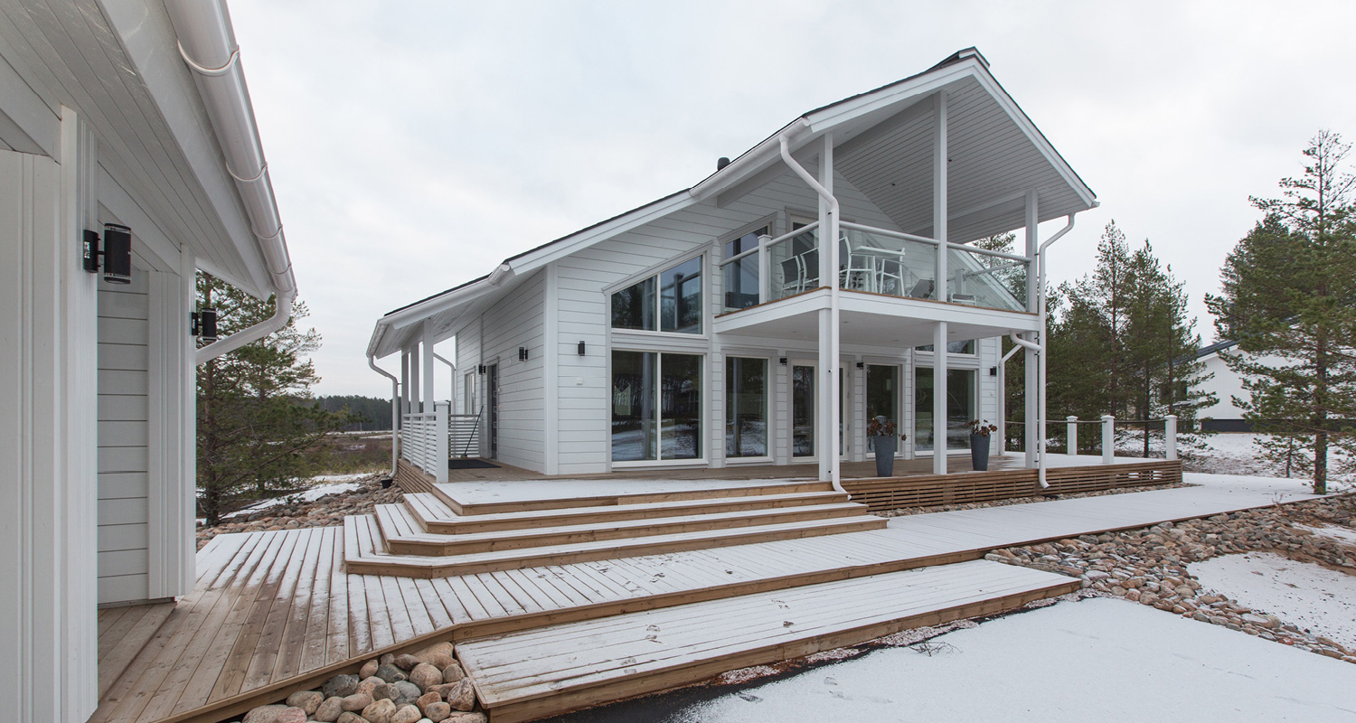 Valkoinen moderni puutalo lumisella terassilla järven rannalla mäntyjen keskellä