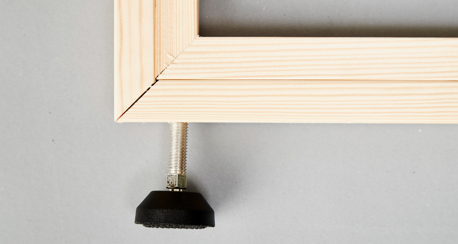 Vaalea puukehyksen kulma ja säädettävä metallijalka mustalla kumipäällä harmaalla taustalla