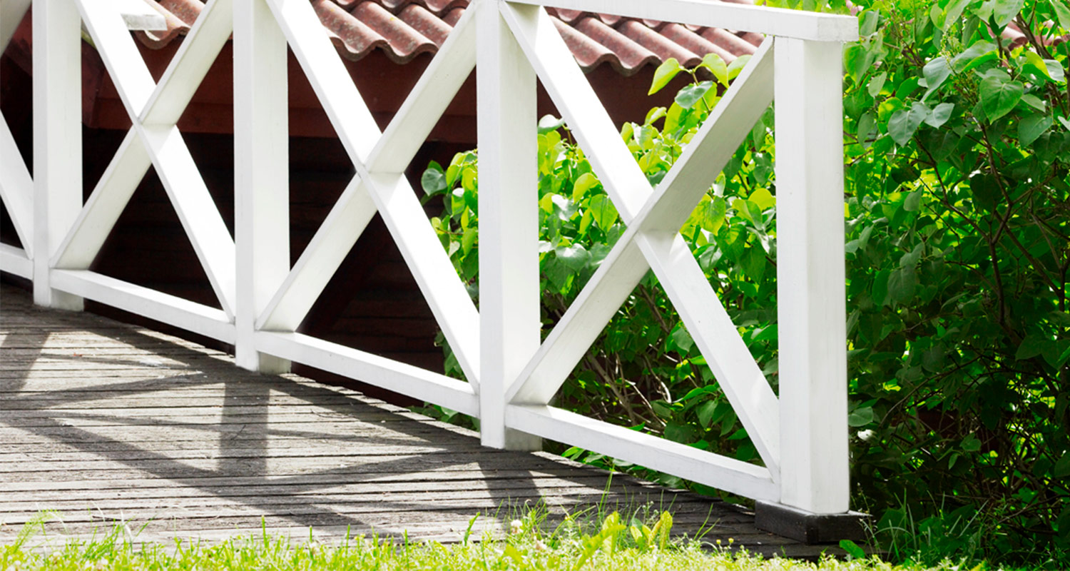 Valkoinen X-mallinen puuaita puisella terassilla, varjoja ja vihreää pensasta taustalla.