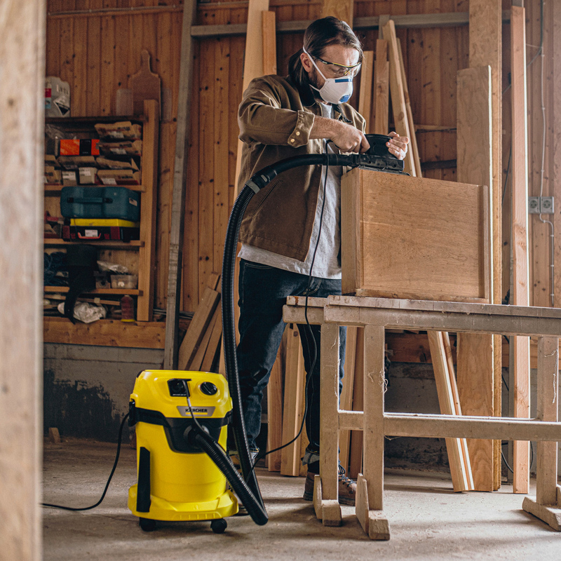 Henkilö imuroi työpajassa puulaatikkoa, keltainen Kärcher-imuri lattialla.