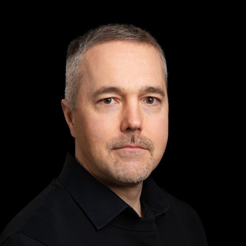 Mies harmaantuvilla hiuksilla mustassa kauluspaidassa tummalla taustalla.