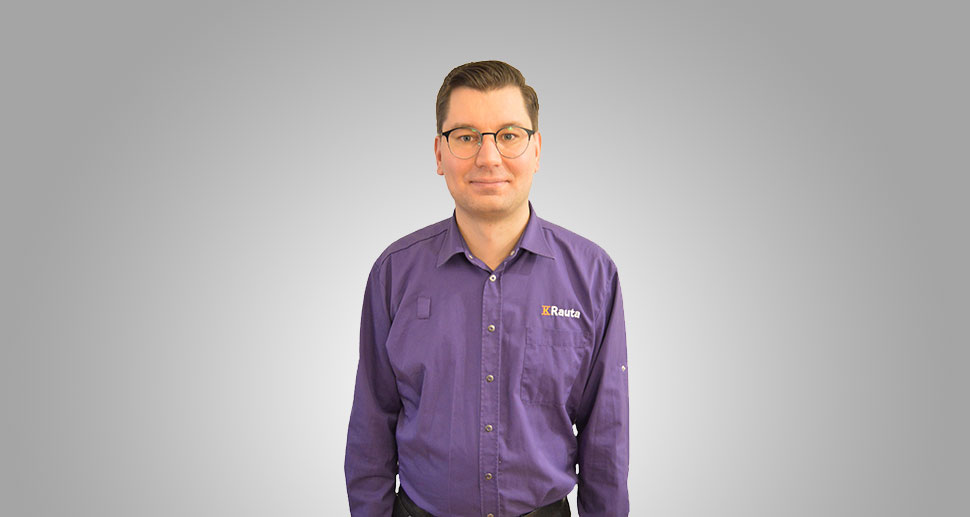 Mies seisoo harmaalla taustalla, yllään violetti K-Rauta-työasu.