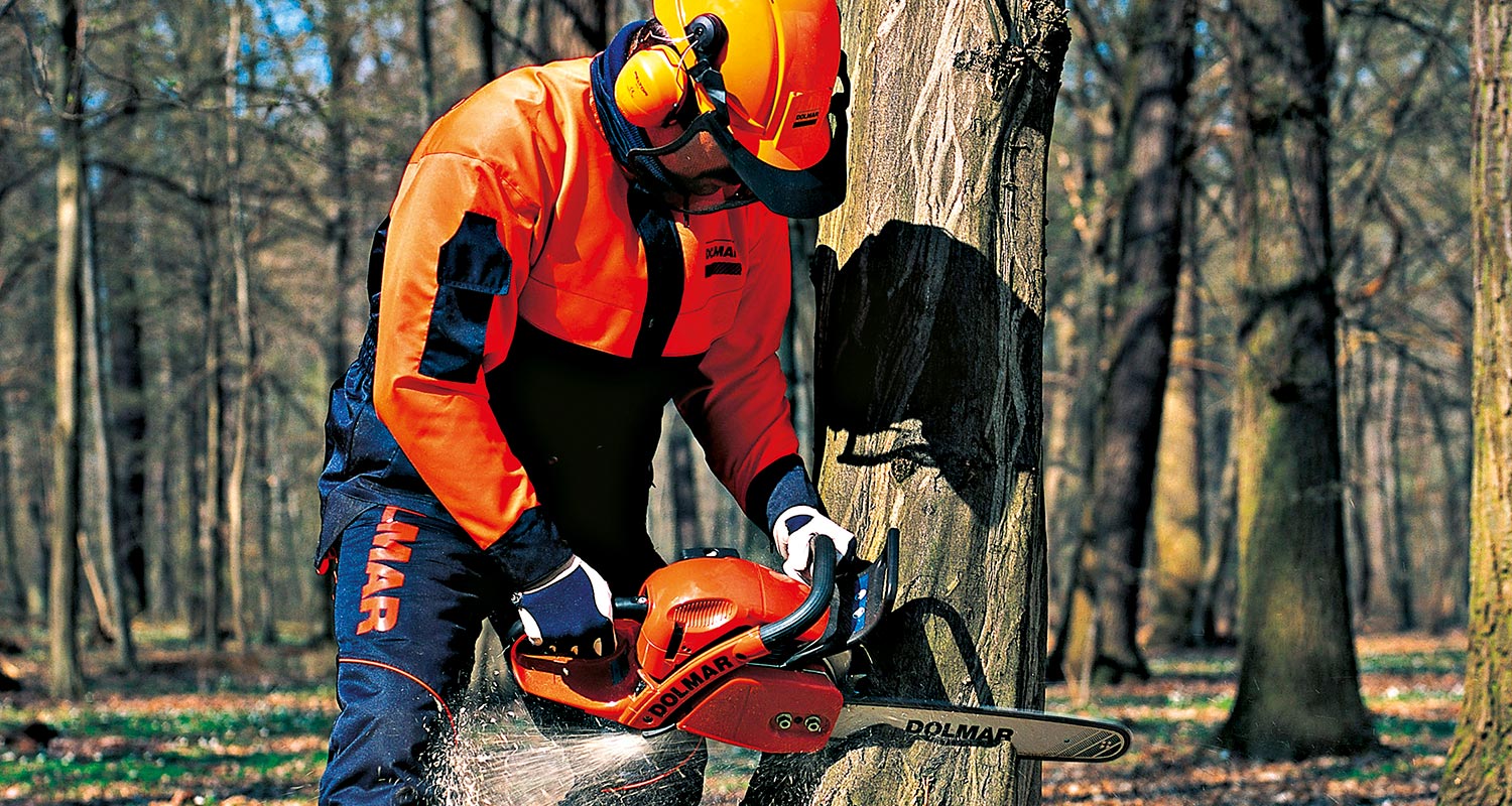 Oranssipukuinen metsuri sahaamassa puunrunkoa oranssilla ketjusahalla vehreässä metsässä.