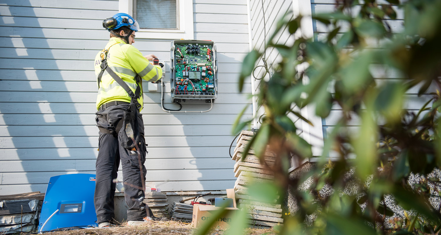 Keltapukuinen asentaja sinisessä kypärässä säätää avoimen sähkötaulun harmaalla puuseinällä