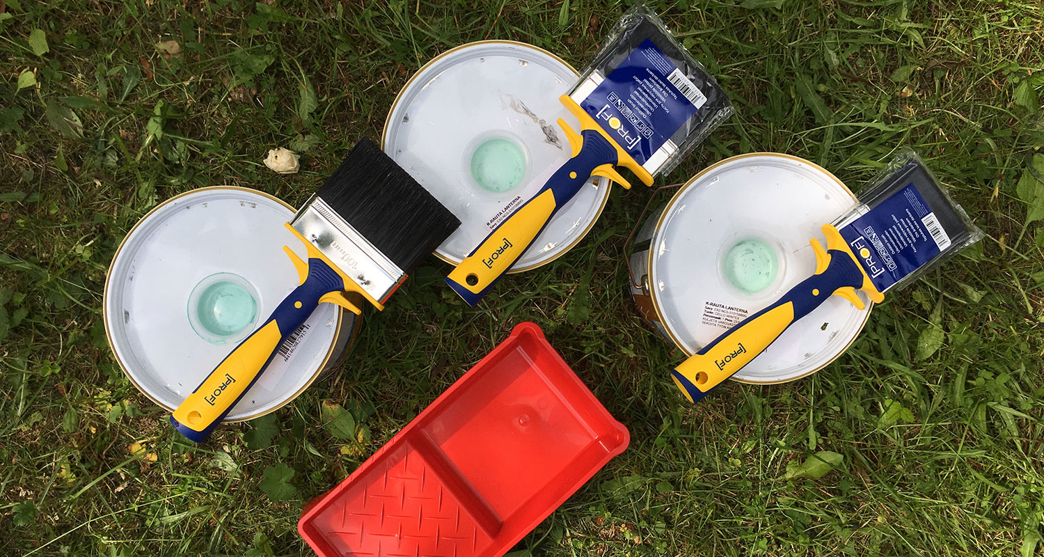 Kolme maalipurkkia valkoisin kansin, kelta-siniset pensselit ja punainen maalauslokero nurmikolla.