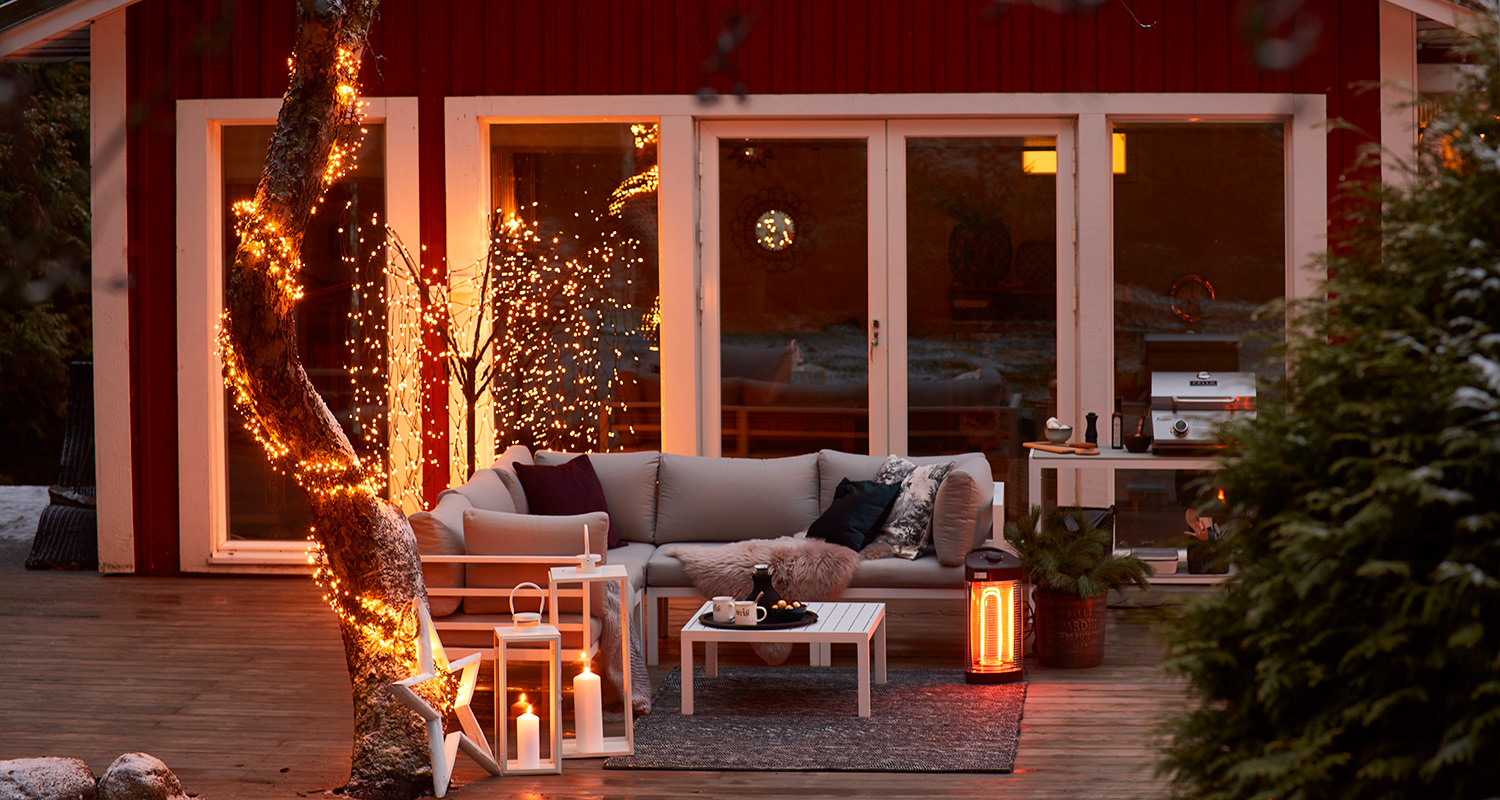 Punaisen mökin terassilla puunrungossa keltaiset valot, valkoinen sohva, lyhdyt ja lämmitin.