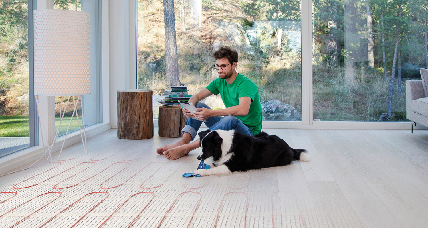 Mies tabletilla vaalealla lattialla, koira leikkii lattialämmityksen päällä, ikkunasta näkyy metsä.