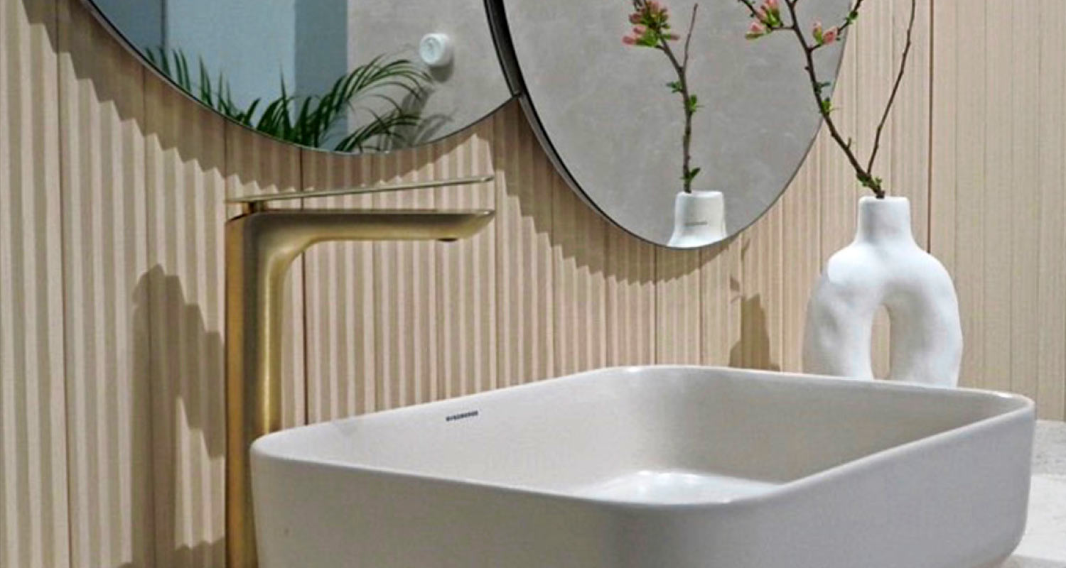 Valkoinen kulmikas pesuallas messinkihanalla, peili, beige paneeliseinä ja kirsikankukkamaljakko