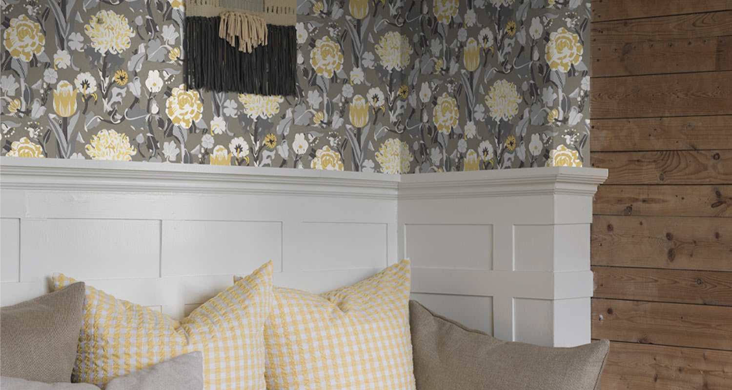 Nurkkakuvassa harmaa-keltainen kukkatapetti, valkoinen paneeli ja kelta-valkoiset ruututyynyt.