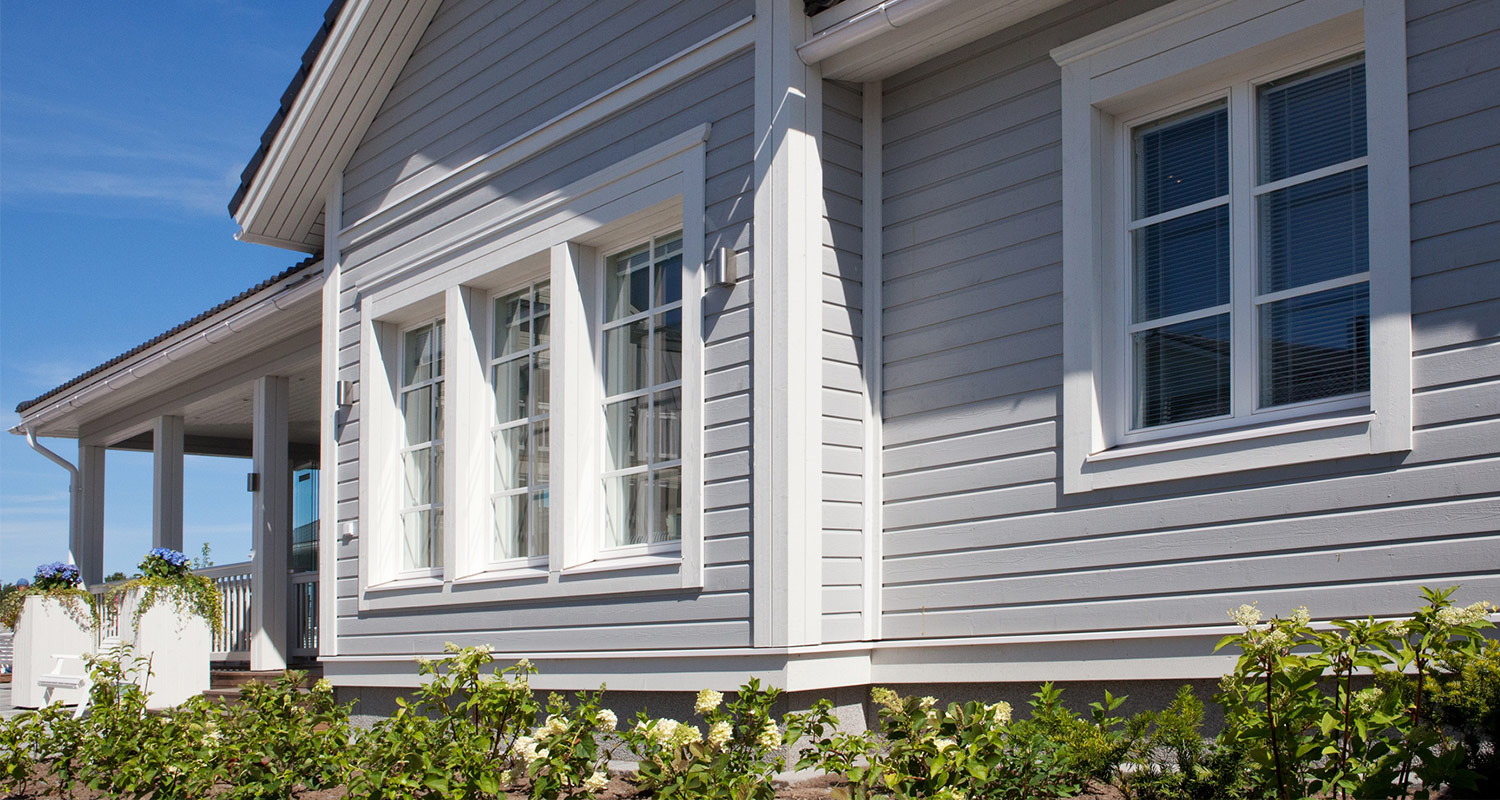 Vaaleanharmaa puutalo valkoisilla ikkunoilla, terassin sinisiä kukkia ja vihreää pensastoa.