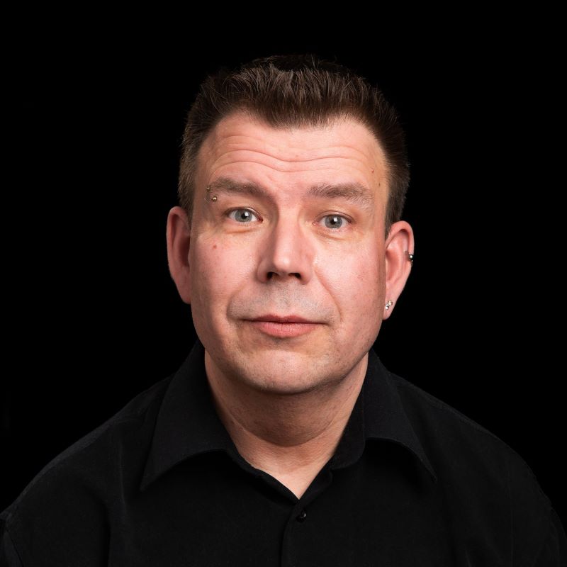 Lähimuotokuva miehestä: lyhyet ruskeat hiukset, korvarenkaat, musta kauluspaita, musta tausta.