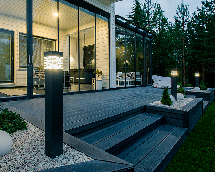 Moderni harmaa terassi mustilla pylväslampuilla lasiseinän vieressä, metsätausta