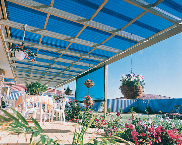 Aurinkoisen terassin sininen katos, valkoiset muovituolit, roikkuvia kukkakoreja ja pinkkejä kukkia