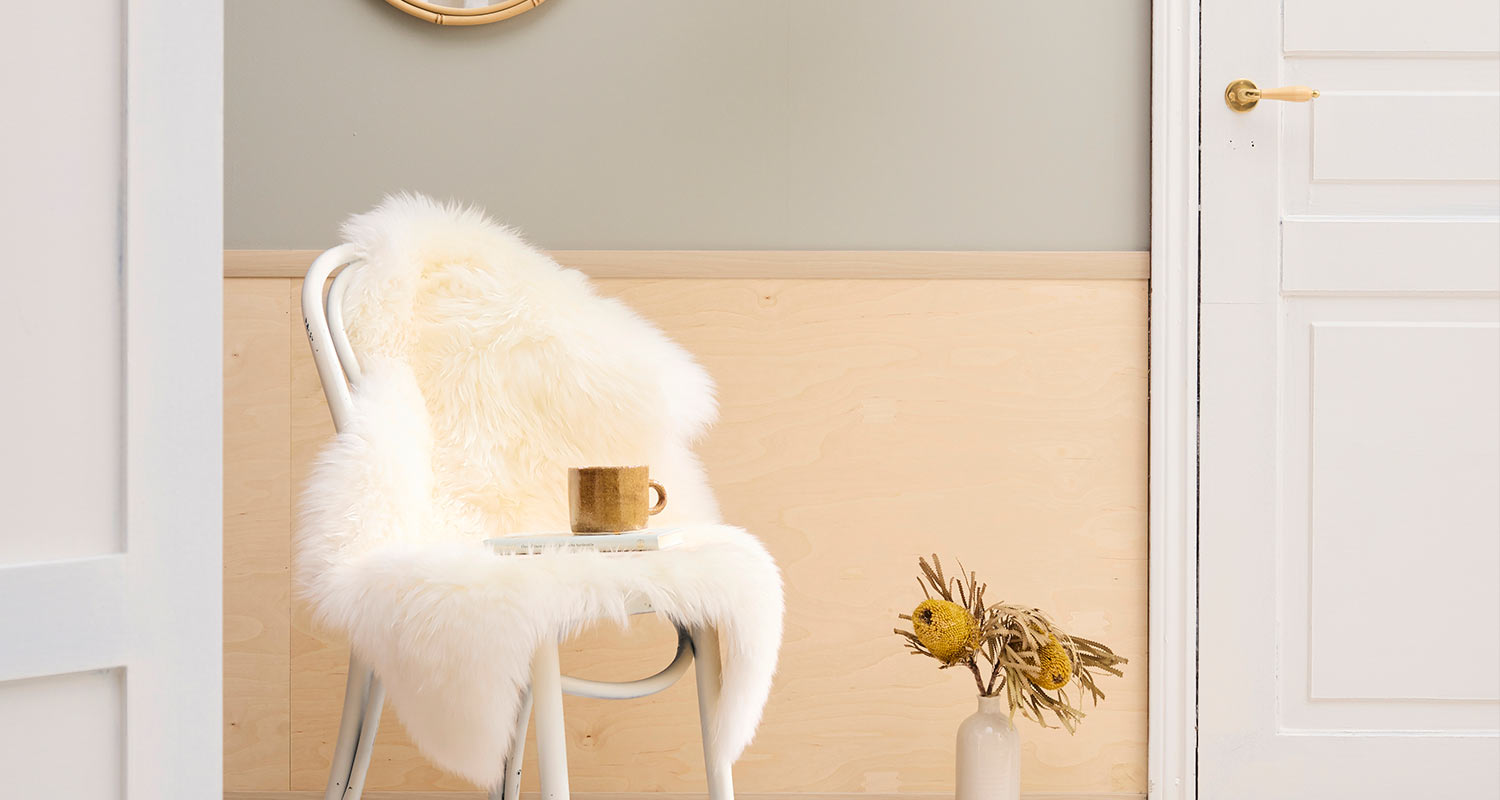 Vaalea sisätila: valkoinen tuoli lampaantaljalla, kahvimuki ja kirja, maljakko kuivakukilla.