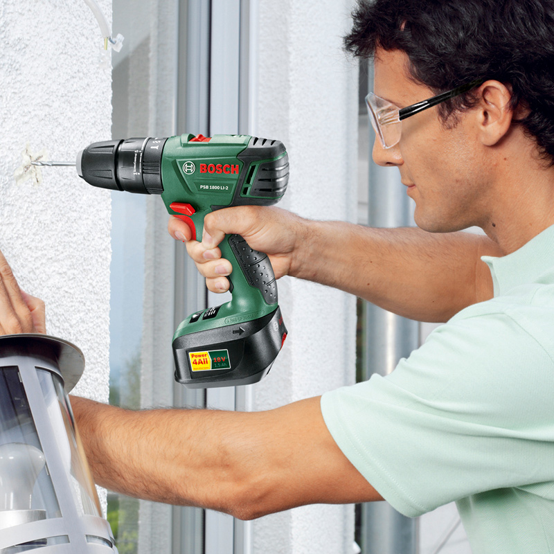 Mies kiinnittää ulkovalaisinta valkoiselle seinälle vihreällä Bosch-akkuporakoneella