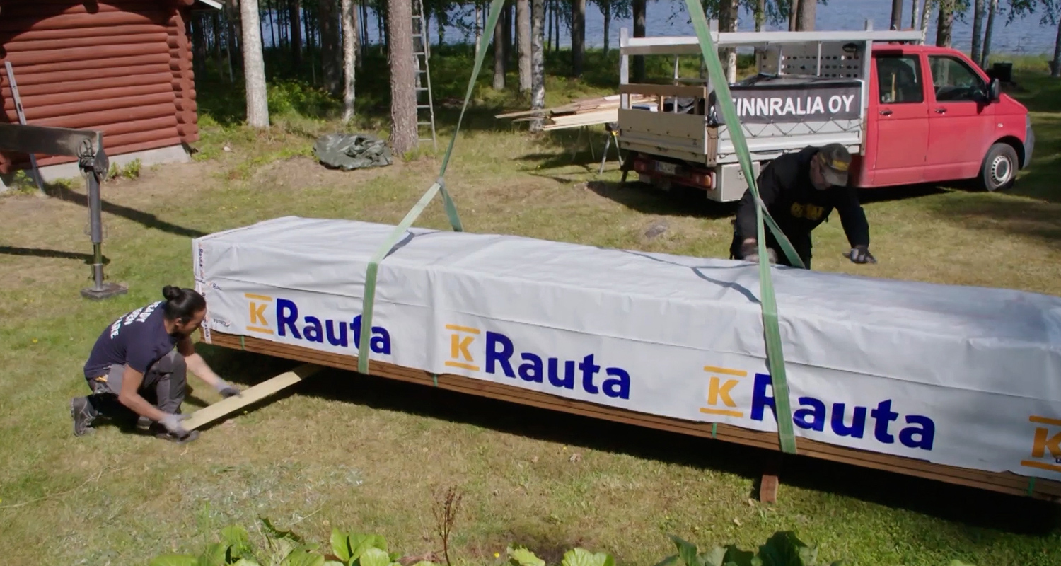 K-Rauta-pakkaus nostetaan nurmella, taustalla punainen pakettiauto ja metsä