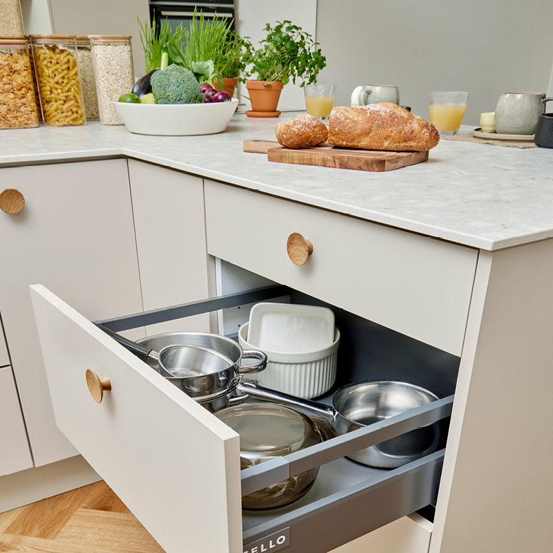 Vaalea keittiölaatikko auki, sisällä metallikattiloita, päällä vaaleaa leipää ja vihanneksia.