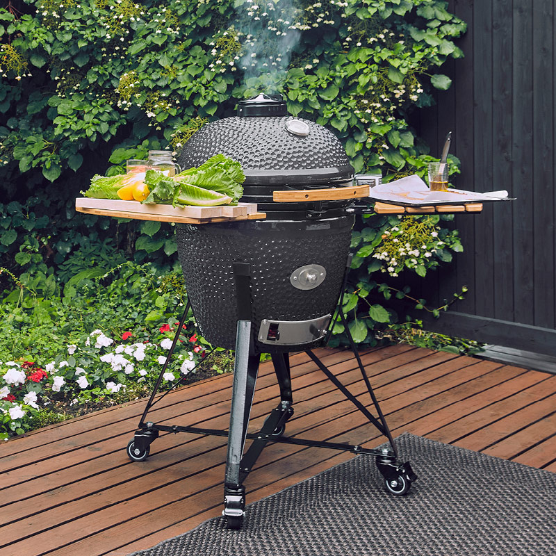 Mustan kamadogrillin sivuhyllyillä salaattia ja keltaista paprikaa, terassilla vihreän pensasaidan edessä.