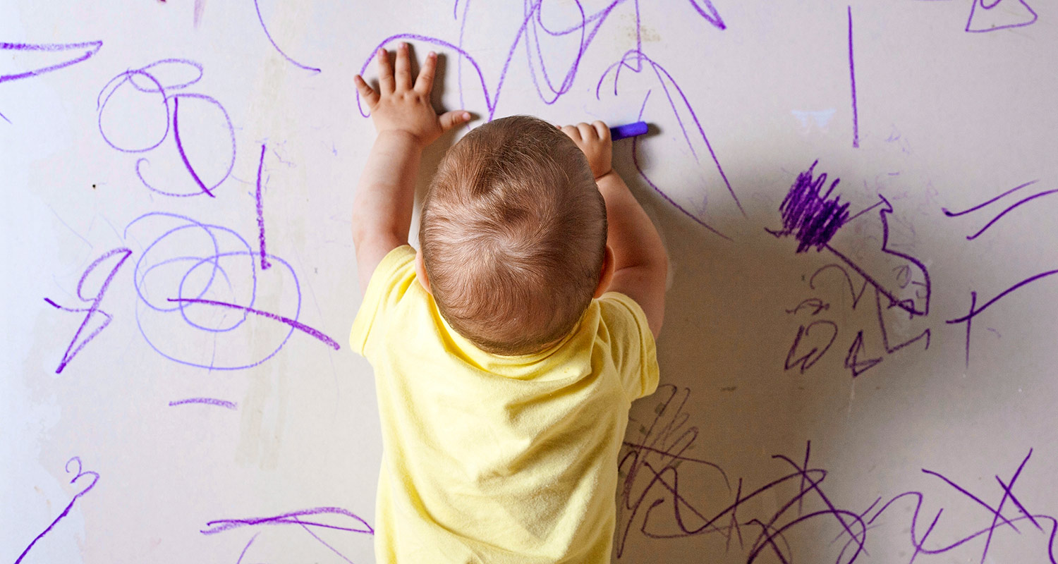 Vauva keltaisessa paidassa piirtää violeteilla tussilla valkoiselle seinälle.