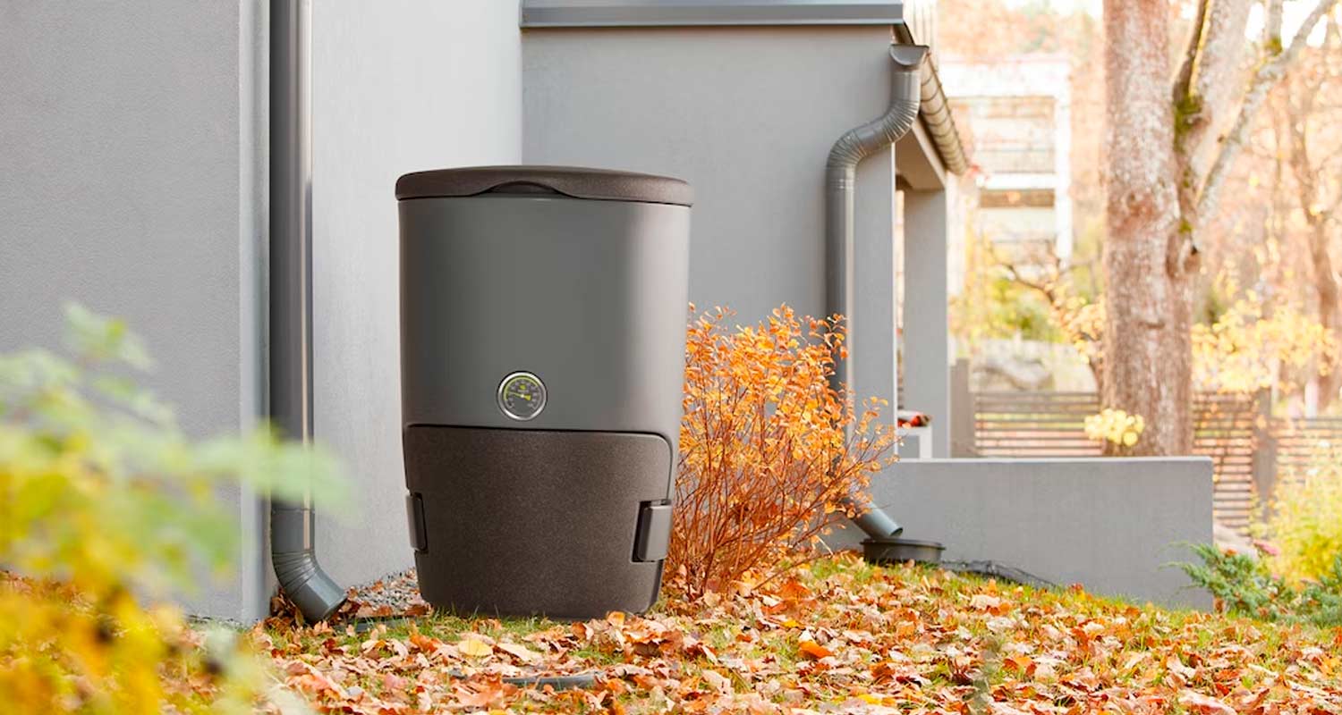 Talon kulmalla harmaa sadevesisäiliö mittarilla, maassa ruskeita lehtiä ja oransseja pensaita.