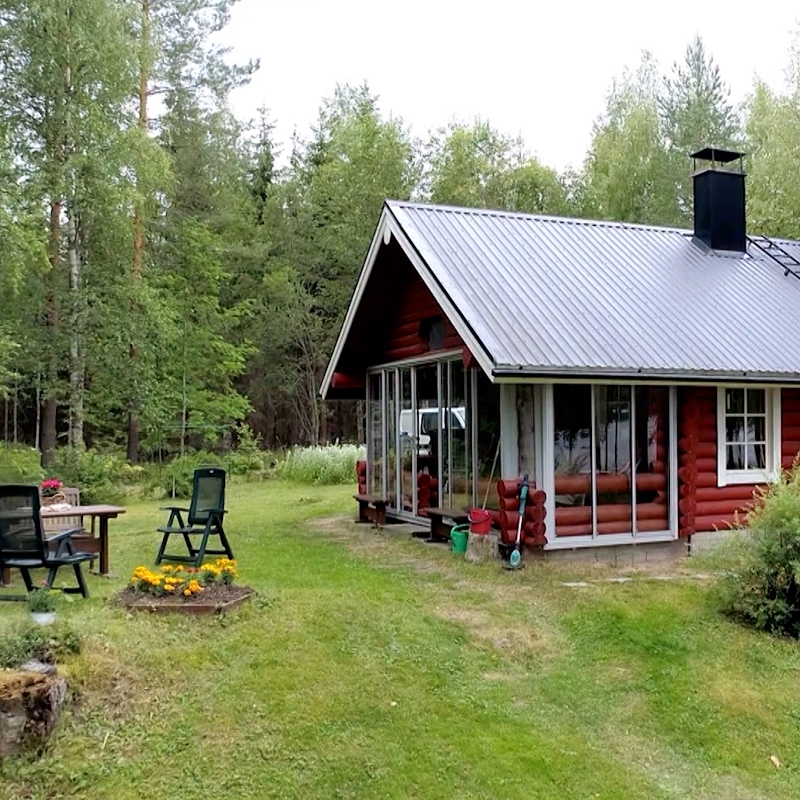 Punainen hirsimökki metallikatolla ja lasitetulla kuistilla metsäaukealla, pihatuolit nurmikolla