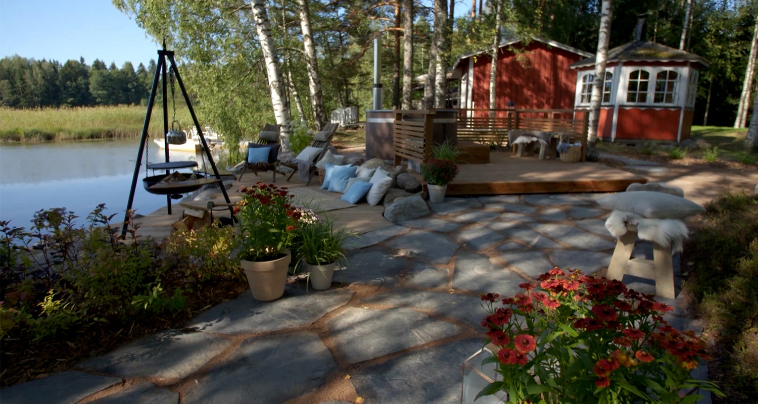 Punainen mökki järven rannalla, koivuja, terassilla tyynyjä, kukkaruukkuja ja grillikolmio.