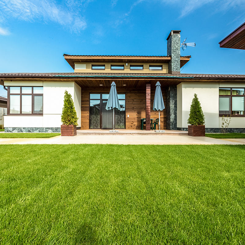 Moderni omakotitalo puupaneelilla, sininen taivas, harmaat aurinkovarjot, vihreä nurmikko.