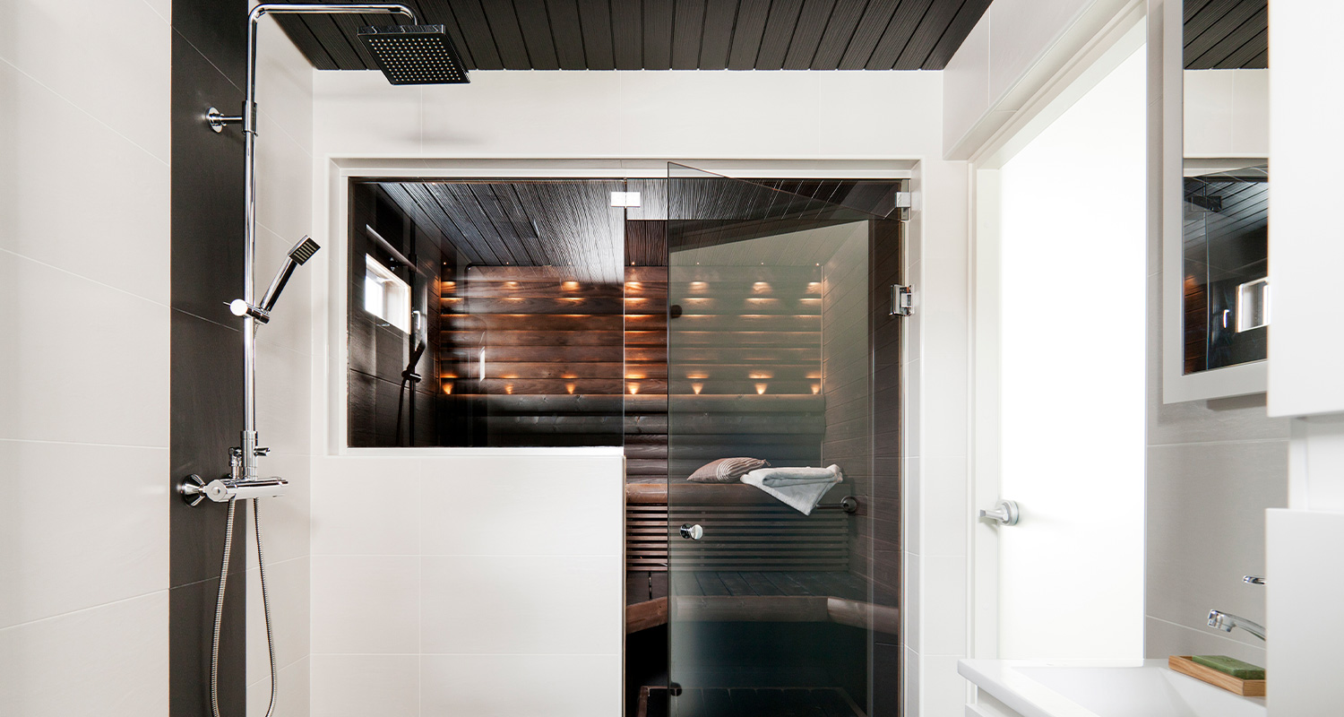 Moderni valkoinen kylpyhuone suihkulla ja lasiseinällä, takana puusauna lämpimillä valoilla.