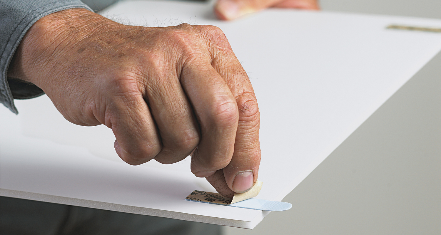 Ikääntyneen käsi repäisee ruskean taustapahvin sinisestä liimatarraosasta valkoisella levyllä.