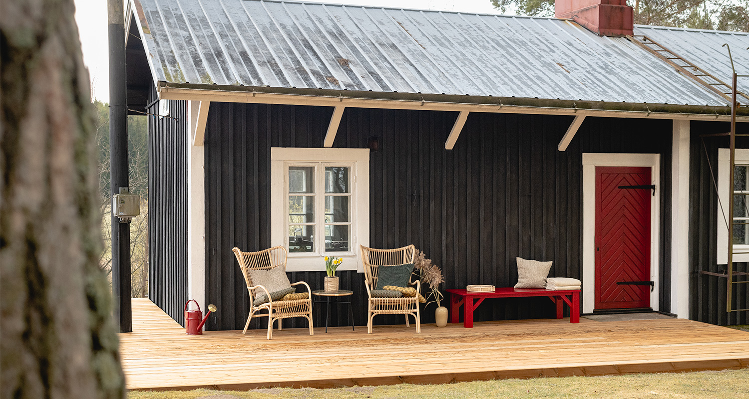 Musta puutalo kuistilla korituolit tyynyin, kukkapöytä, punainen penkki ja ovi