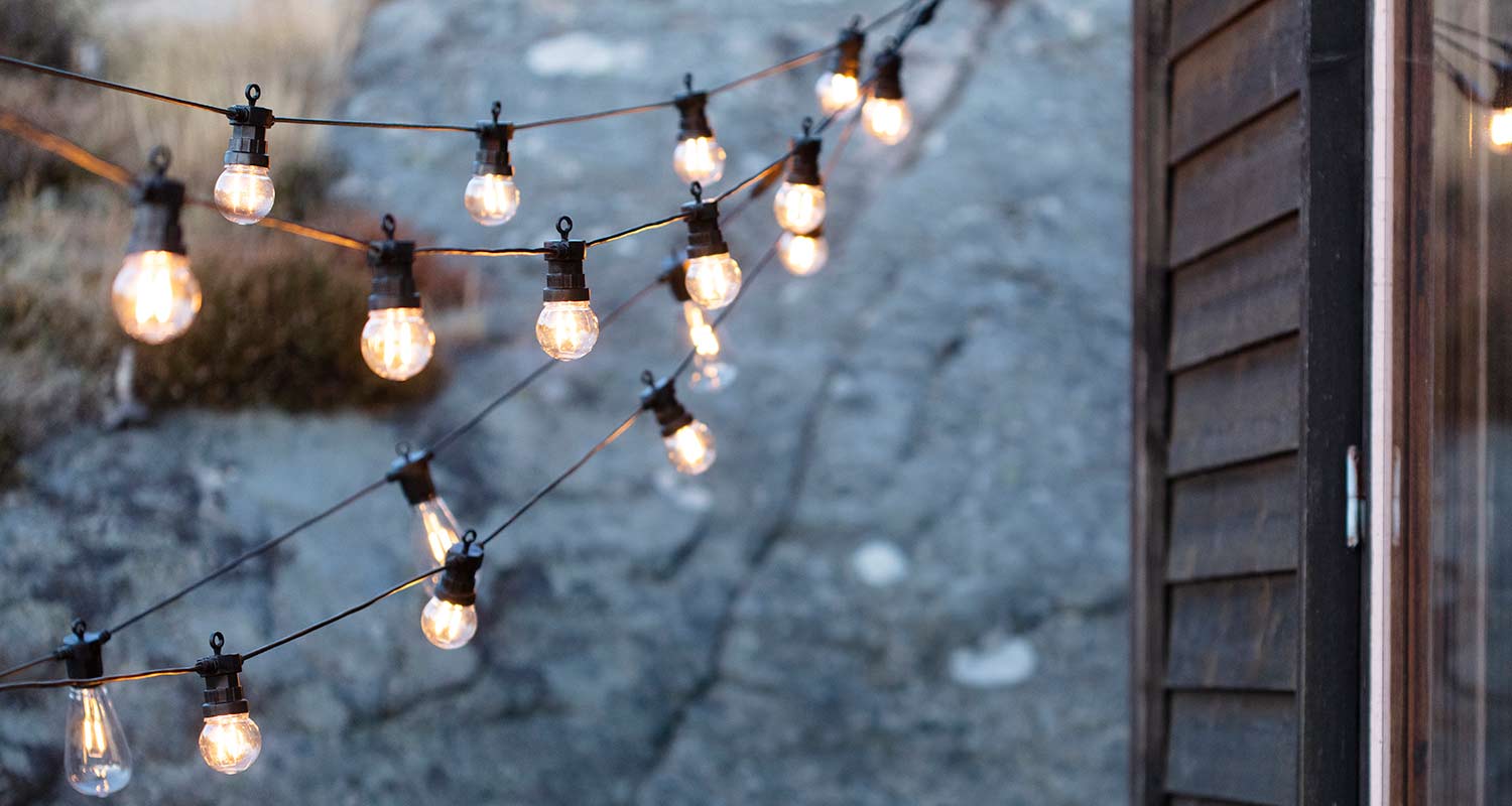 Lämpimät valopallot roikkuvat kolmessa rivissä naruissa kiven ja tumman puutalon edustalla.
