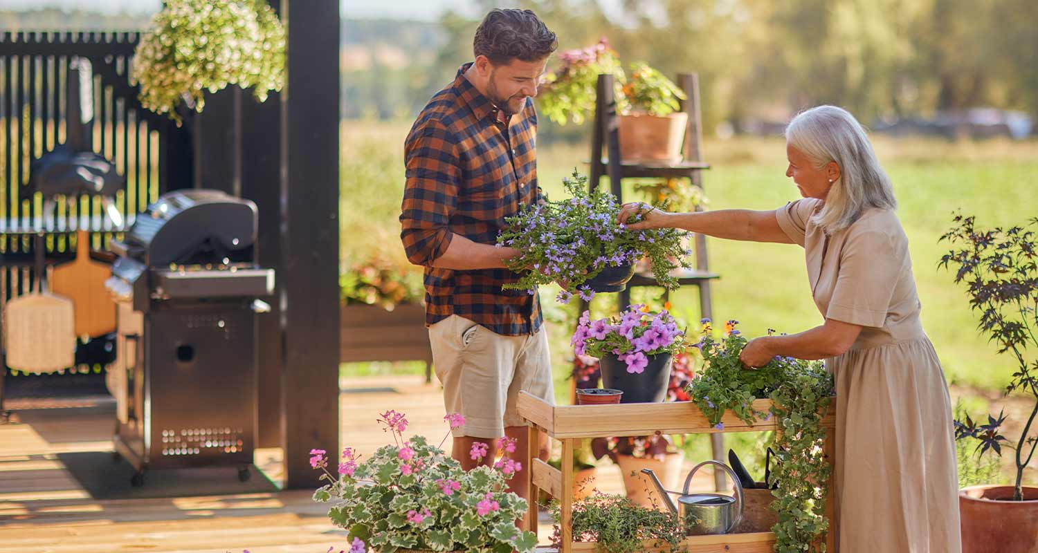 Kesäinen terassi, nainen ja mies istuttavat violetin- ja pinkkikukkia puupöydällä.