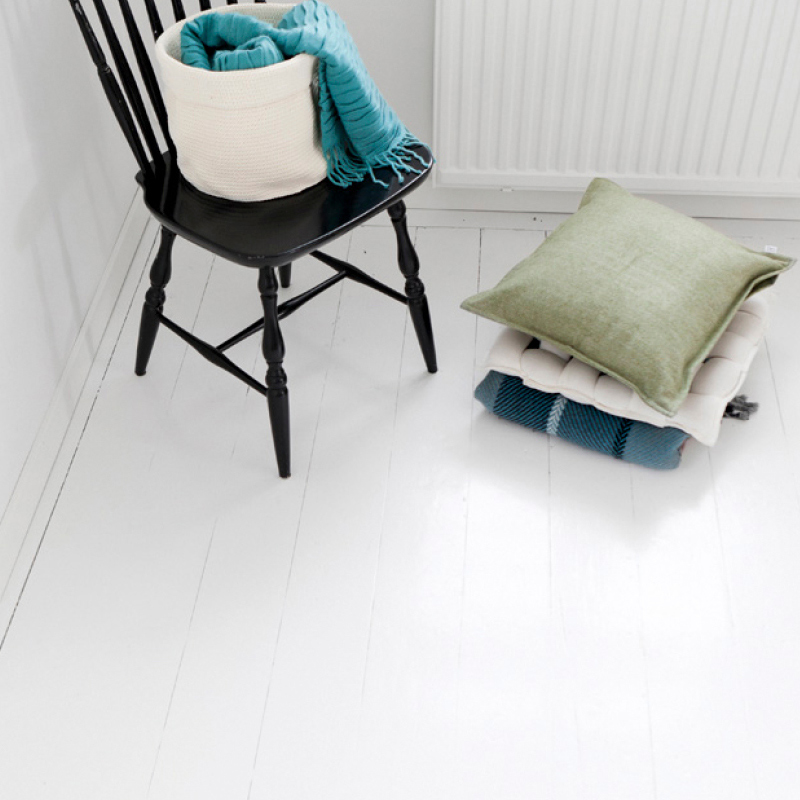 Kuvassa valkoiseksi maalattu lattia. Lattialla on musta tuoli ja muutama tyyny. Tuolin päällä viltti.