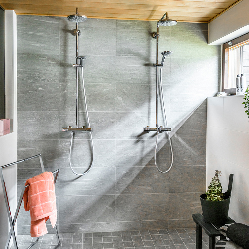 Kaksi hopeista suihkupäätä harmaatiilisessä suihkutilassa, vaalea puukatto, pinkki pyyhe ja huonekasvi