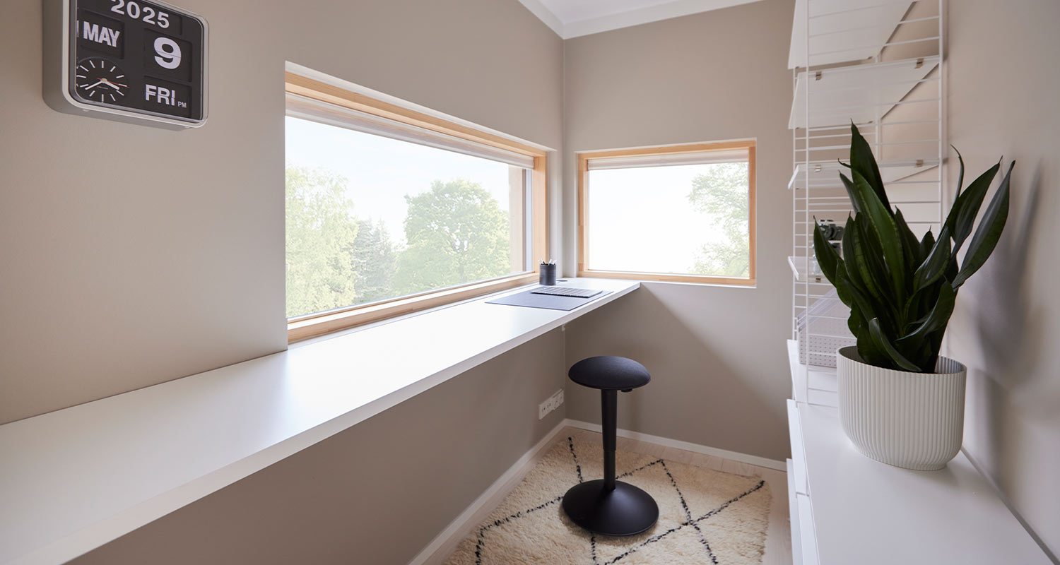 Beigessä nurkkahuoneessa pitkä valkoinen pöytä ikkunan edessä, musta jakkara ja viherkasvi.