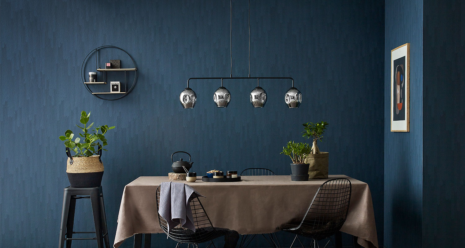 Syvän sininen ruokailutila, ruskea pöytäliina, mustat tuolit, lasivalaisimet ja viherkasveja.