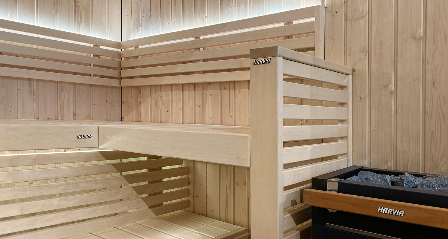 Valoisa kuusipanelinen sauna, L-mallinen laude ja Harvia-kiuas mustine kivineen