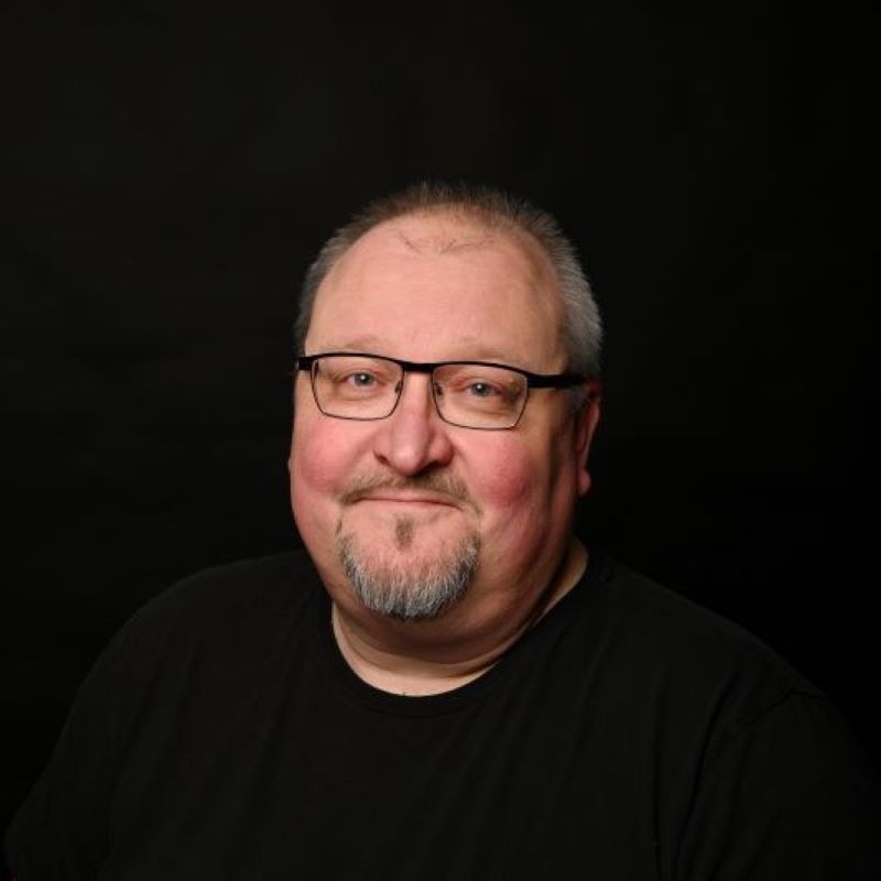 Harmaahiuksinen mies mustassa t-paidassa tummassa studiotaustassa.