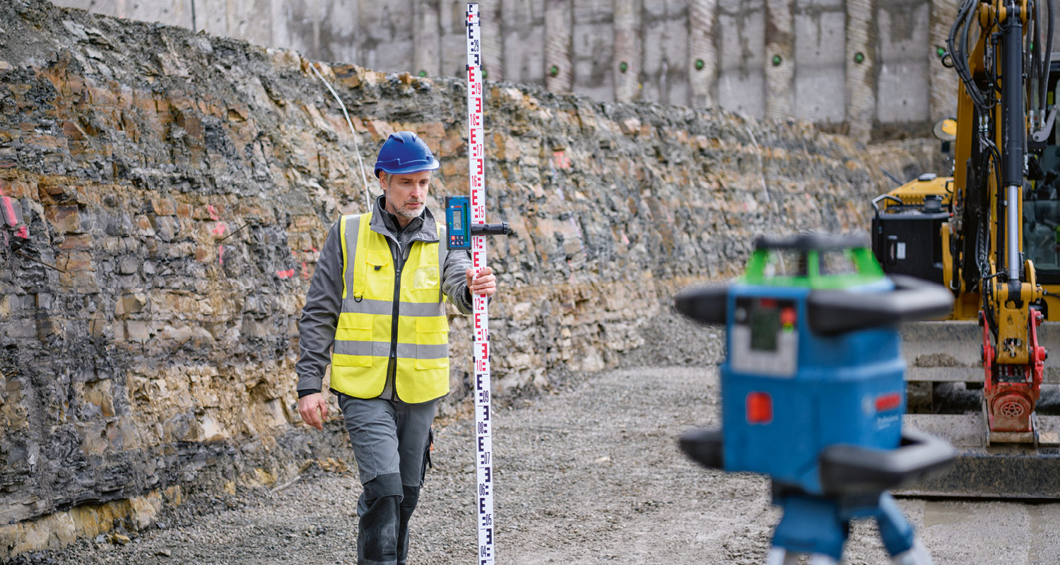 Kaivutyömaalla kypäräpäinen keltaliivinen mittamies pitää mittakeppiä, etualalla sininen laserlaite