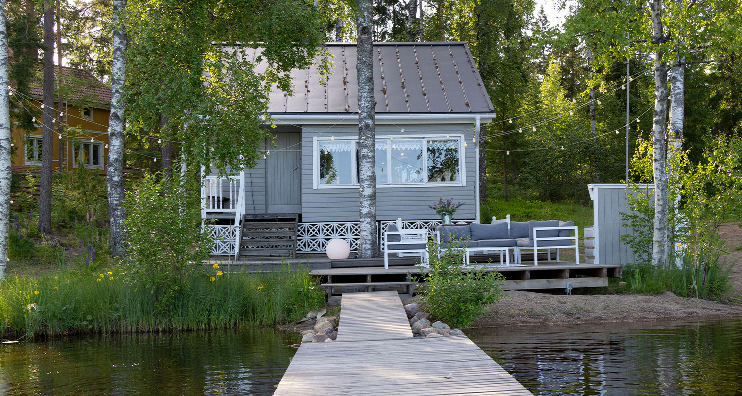 Harmaa kesämökki koivikon reunassa, edessä laituri järveen ja valkoinen sohvaryhmä.