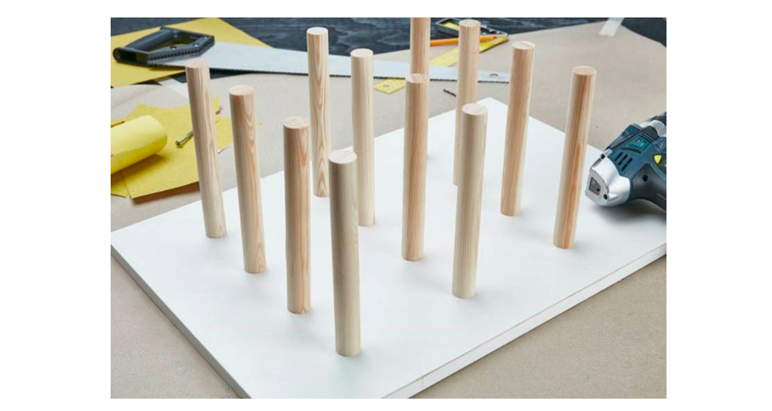 Valkoisella levyllä riveissä pystysuoria puisia tappeja, vieressä porakone ja hiekkapaperi.