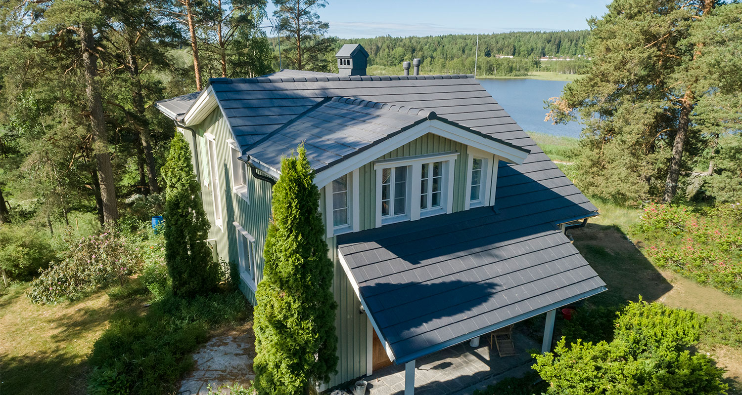 Vaaleanvihreä puutalo tummanharmaalla katolla mäntymetsän reunassa järven rannalla