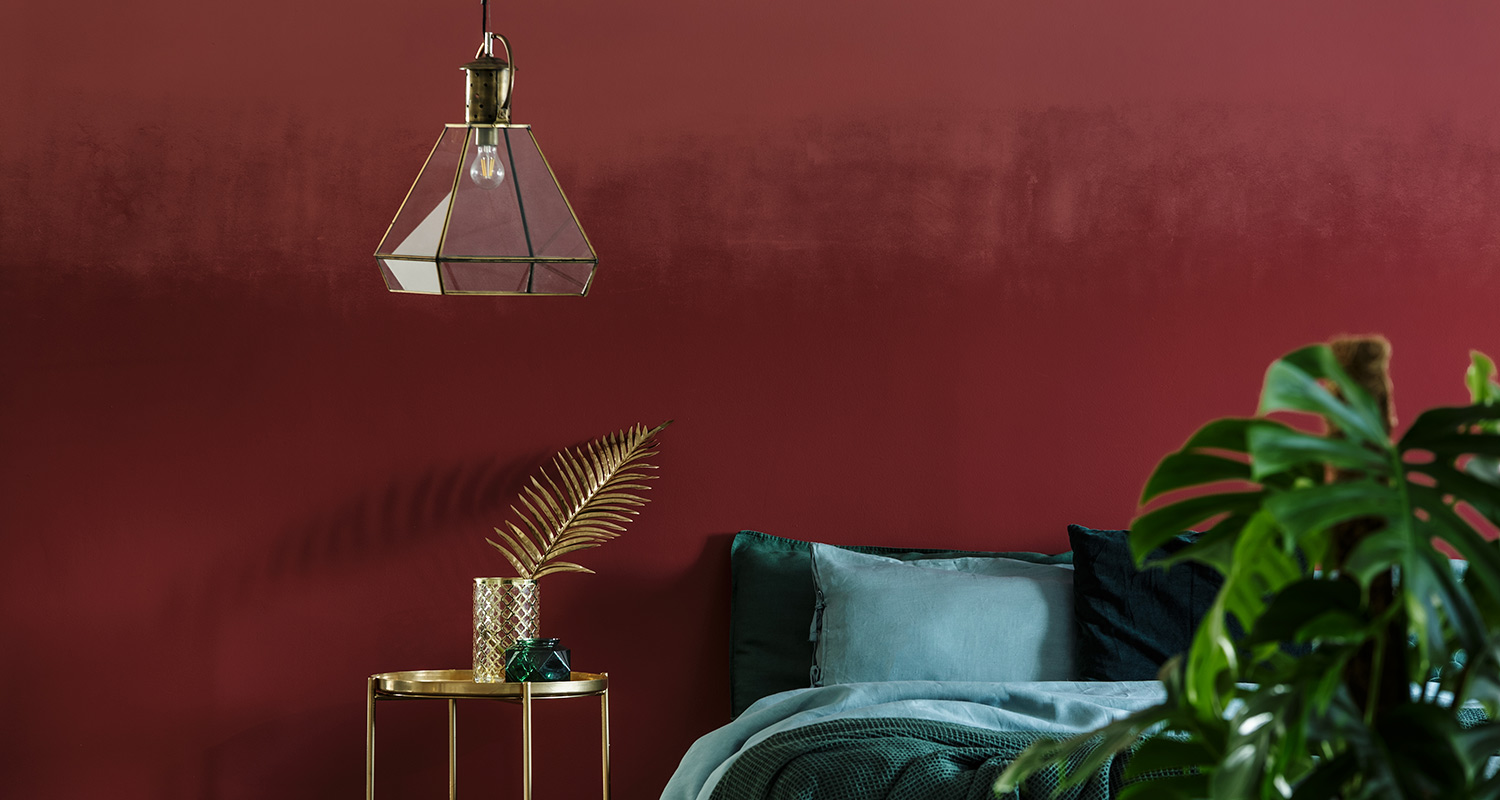 Punainen seinä, lasinen riippuvalaisin, messinkipöytä kultalehdellä, turkoosi sänky ja viherkasvi.