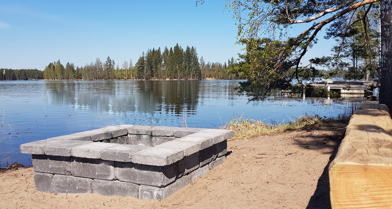 Harmaa kivinuotiopaikka hiekkaisella rannalla, sininen järvi heijastaa havumetsää taustalla