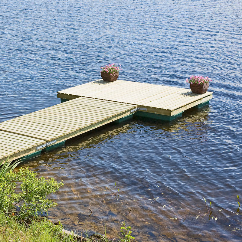 Puisella laiturilla kaksi ruukkua vaaleanpunaisia kukkia, sininen järvi ja vihreä ranta.